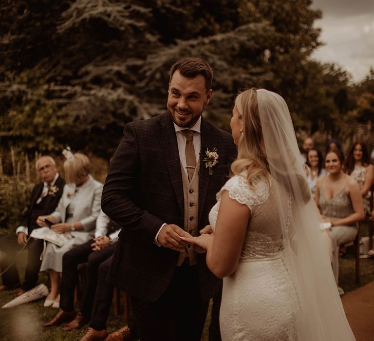 Bride in a lace cap sleeve wedding dress and cathedral length veil exchanging vows with her husband at Dewsall Court