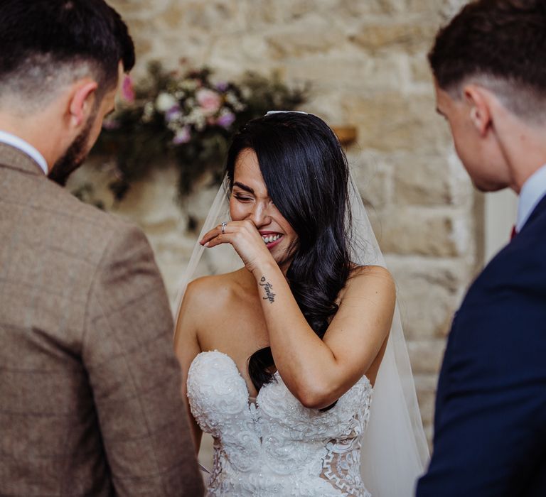 Bride in lace wedding dress with sweetheart neckline laughs and she stands with groom in brown check suit during wedding ceremony