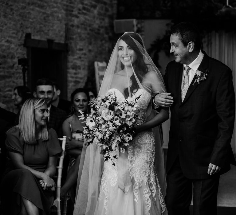 Bride in lace and tulle wedding dress with sweetheart neckline and veil over her face holding multicoloured bridal bouquet walks down the aisle with her father during stone barn wedding ceremony