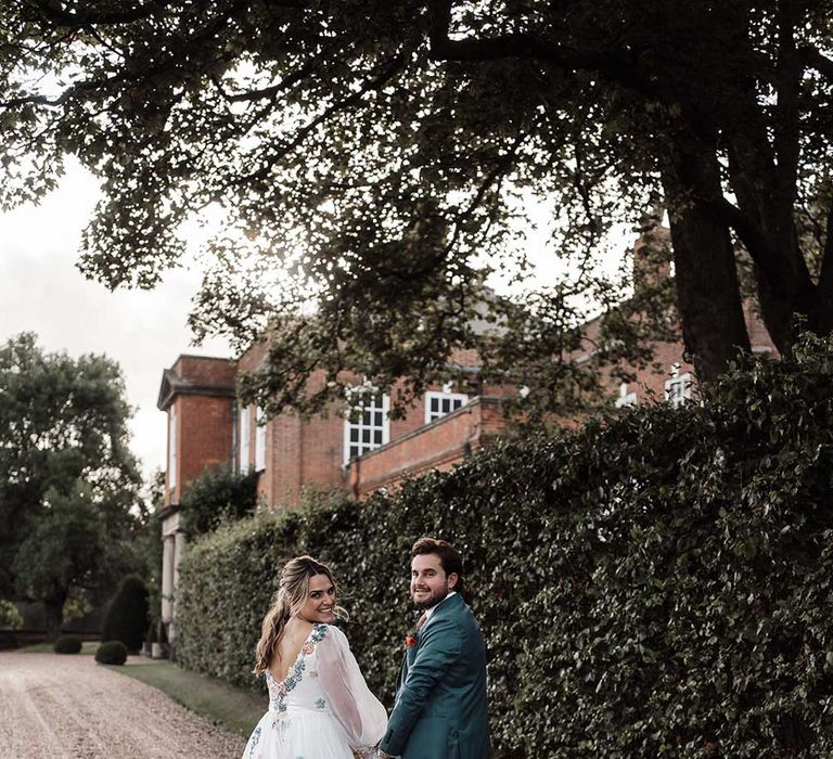 Smiling bride in bespoke Victoria Sanders wedding dress with train and floral embroidery walks down drive holding hands with groom in blue suit