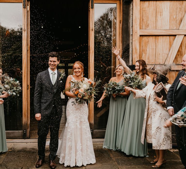 Guests throw confetti over bride in lace and tulle wedding dress holding mixed bridal bouquet and groom in dark suit and blue tie after barn wedding ceremony