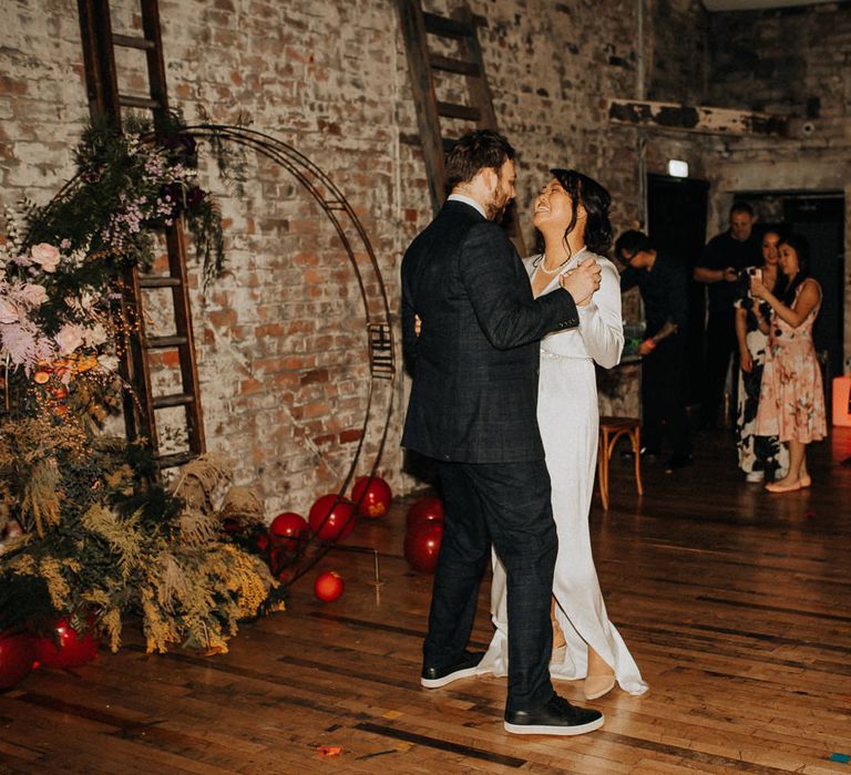 Bride in white long sleeve satin dress and wedges dances with groom in check suit at evening wedding reception