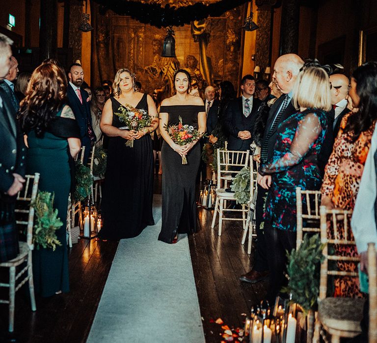 Bridesmaids in black dresses in various styles walk down the aisle together holding bright and colourful bouquets