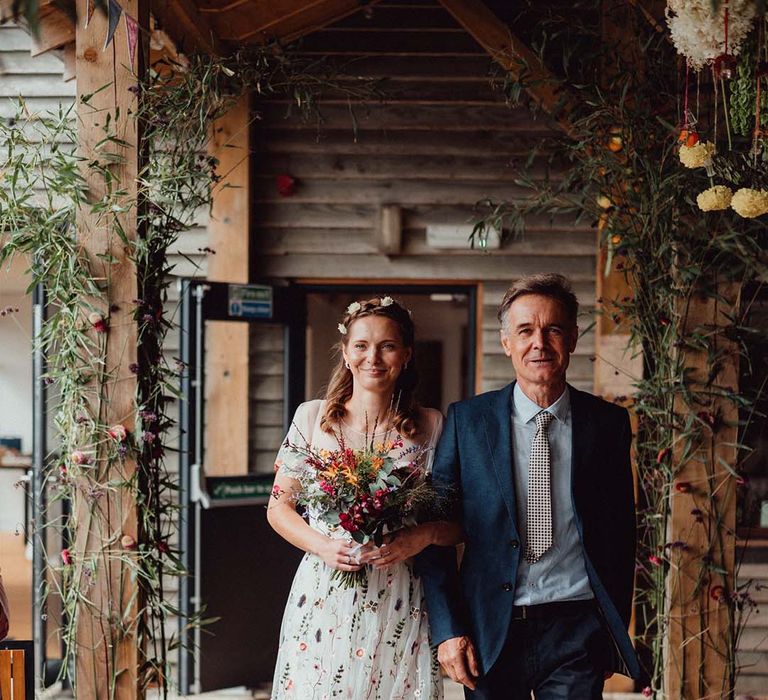 Father of the bride in blue suit and checkered tie walks the bride down the aisle in floral dress