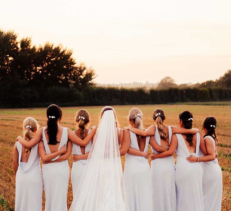 Bride in lace wedding dress with long train and veil with white flower headband with bridesmaids in grey draped open back dresses with white flowers in their hair