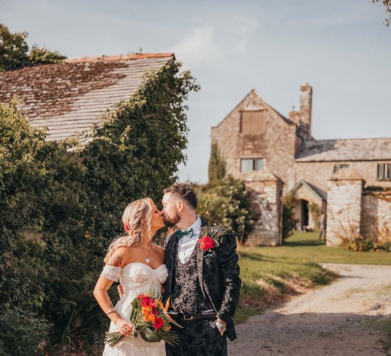 Bride and groom share a kiss outside of The Ash Barton wedding venue in their unique outfits and bright colourful bouquet 