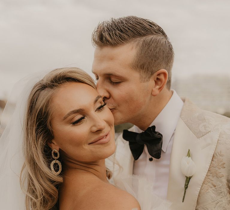 Groom kisses bride on the cheek in gold tuxedo and white tulip buttonhole with bride wearing veil and gold diamanté earrings