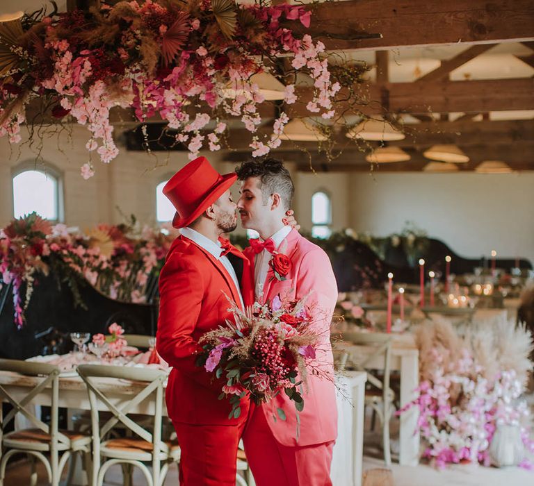 Groom in a red suit and top hat holding a pink flower and dried grass bouquet kissing his partner in a pink suit at their rustic luxe wedding reception at Prestwold Hall Barn 