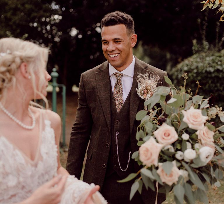 Groom in brown three piece wedding suit carrying the bridal bouquet smiles at the bride in lace wedding dress