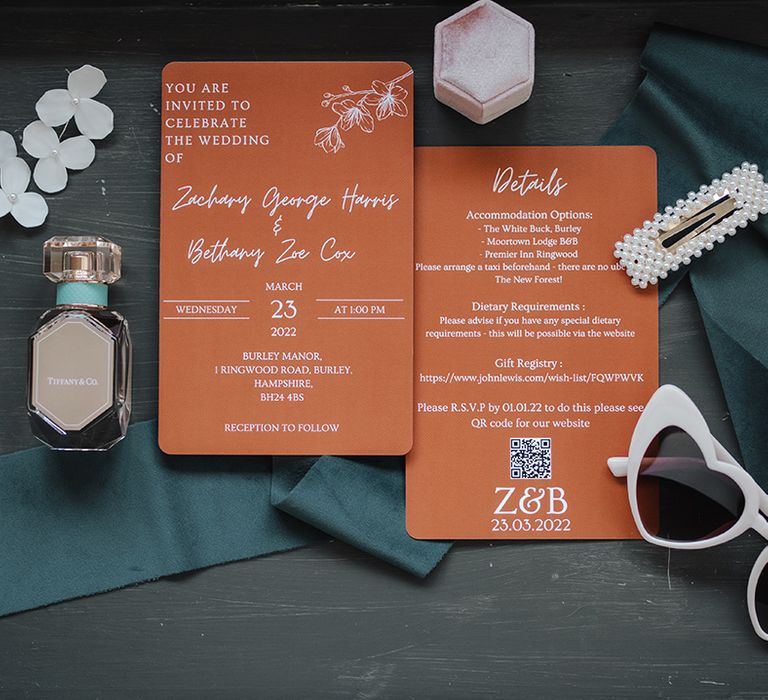 Orange rust coloured wedding invitations with bridal accessories including pearl hair clip, white flower earrings, pink velvet ring box, cat sunglasses and Tiffany perfume