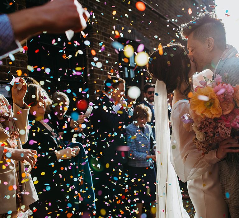 Bride and groom share a kiss as their guests throw multi-coloured confetti over them with bride holding spring bouquet with orange giant poppy 