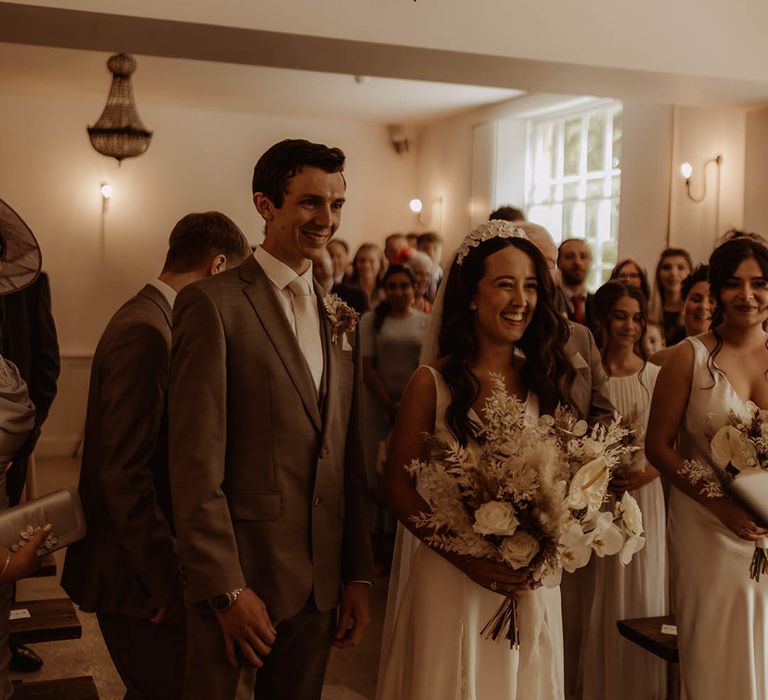 Bride in flower bridal headband smiles with the groom in neutral three piece suit for their boho luxe wedding 