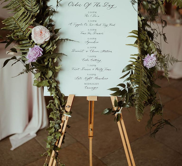 Order of the day wedding sign covered in green foliage, fern leaves and pink peonies 