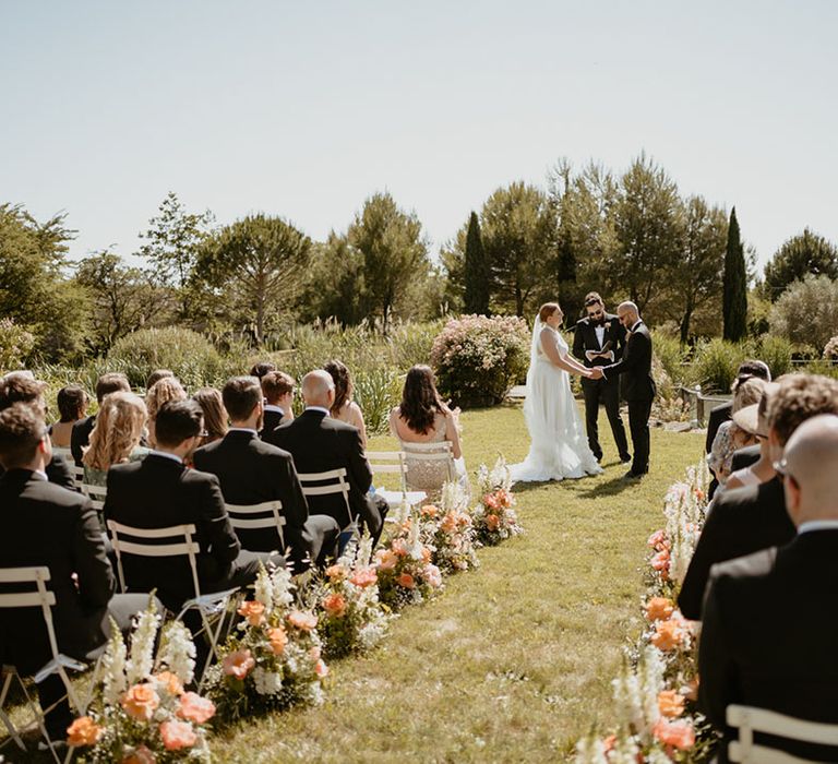 Outdoor wedding ceremony in France with white chairs and aisle lined with peony floral arrangements as wedding guests watch bride & groom marry