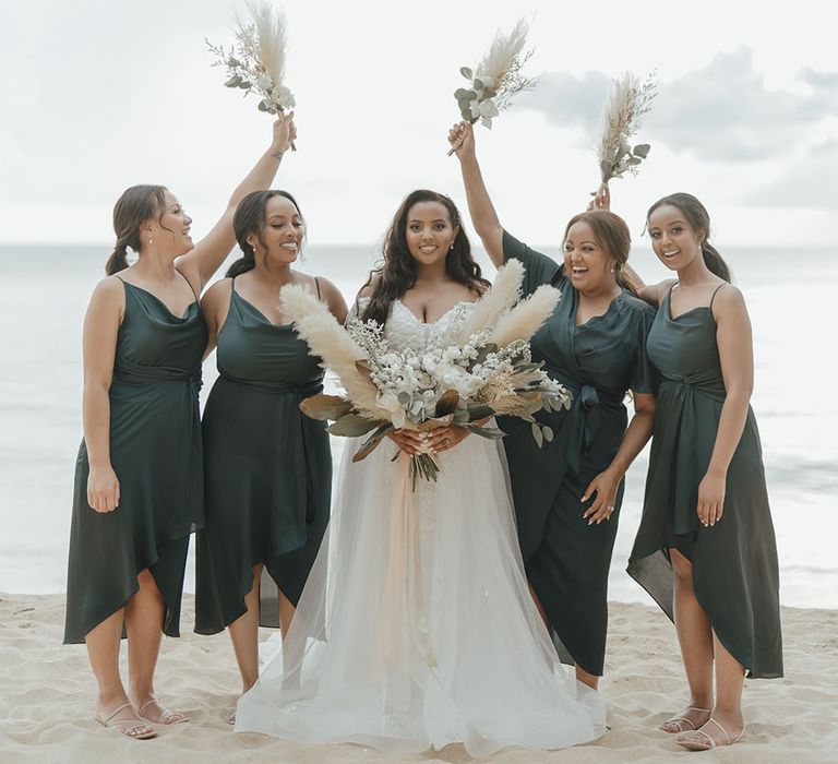 Bride holds pampas grass bouquet and stands with bridesmaids in different styled green satin dresses who hold small pampas grass bouquets