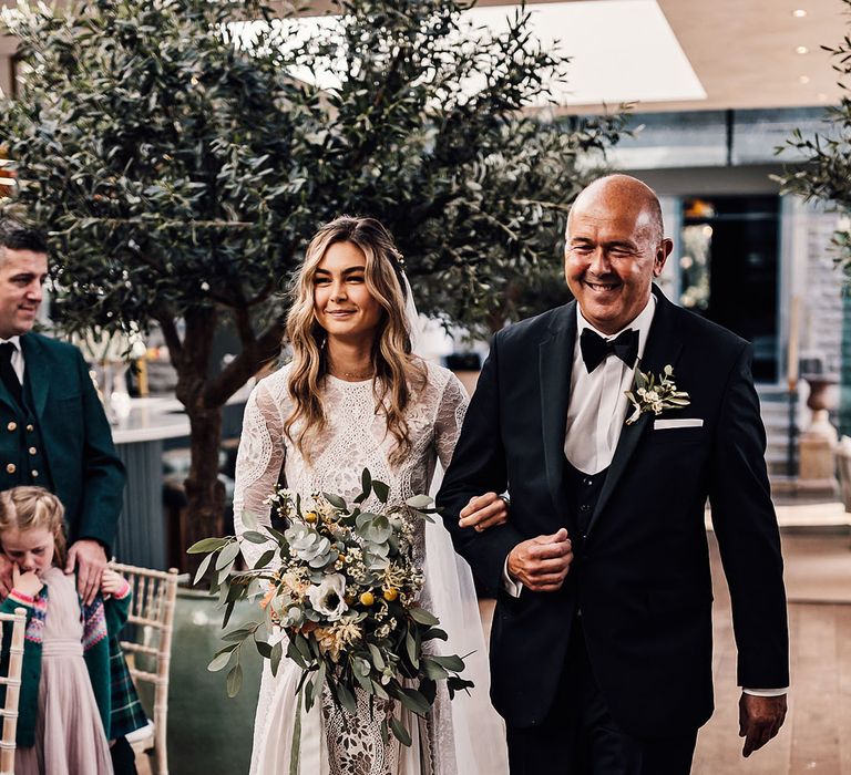 Father of the bride in black tie walks the bride down the aisle carrying a yellow and white bouquet with eucalyptus leaves 