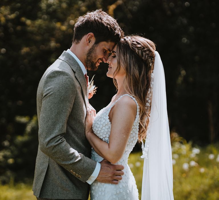 Bride in sleeveless Made With Love Bridal dress with white embellishments with matching hair ribbon and groom in grey suit embracing at The Green Cornwall