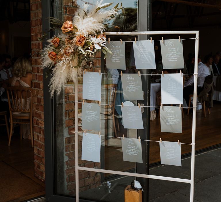 White framed table chart with dried flower and burnt orange and toffee floral arrangement on the corner with minimalist, neutral sheets of paper displaying table information held by small clips 