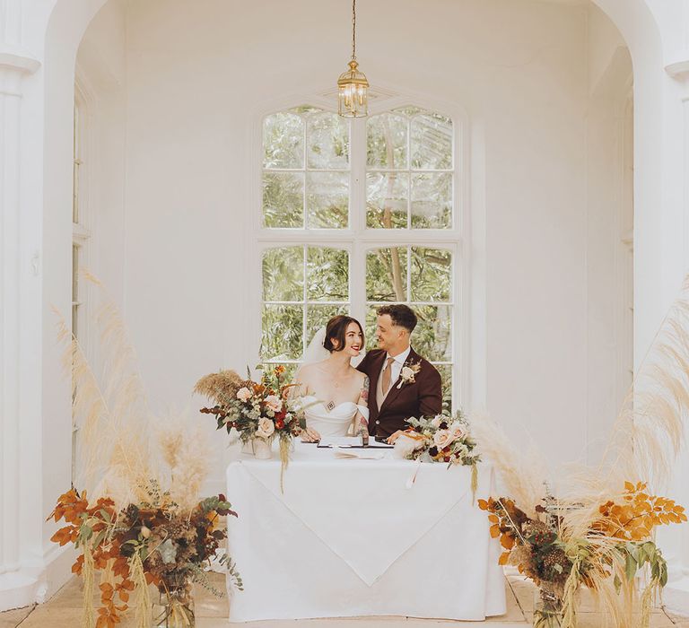 Bride & groom sit at table and sign marriage certificate surrounded by pampas grass floral installations with Autumnal colour scheme