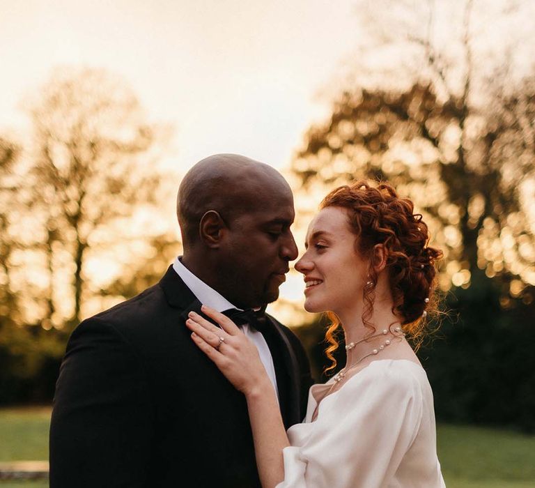 Bride in quarter length sleeve satin wedding dress and groom in black tux embracing at golden hour on the grounds of Pepper Arden Hall