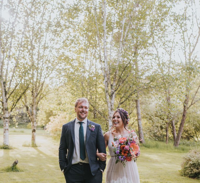 Bride carries colourful floral bouquet as she walks alongside her groom wearing striped suit and colourful floral buttonhole 