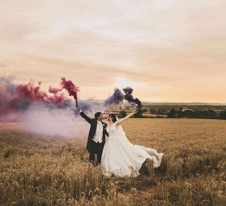 Creative wedding photography showing a Bride and Groom releasing pink and purple smoke bombs in a field