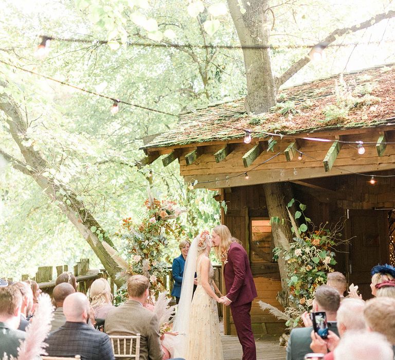 Groom in maroon suit kisses his bride outdoors at the Alnwick Garden Treehouse 