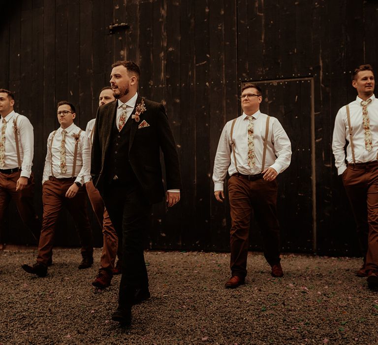 Groom in dark suit with flower patterned tie and pocket square and dried flower boutonniere walking with groomsmen in matching brown trousers, light brown suspenders and flower patterned ties