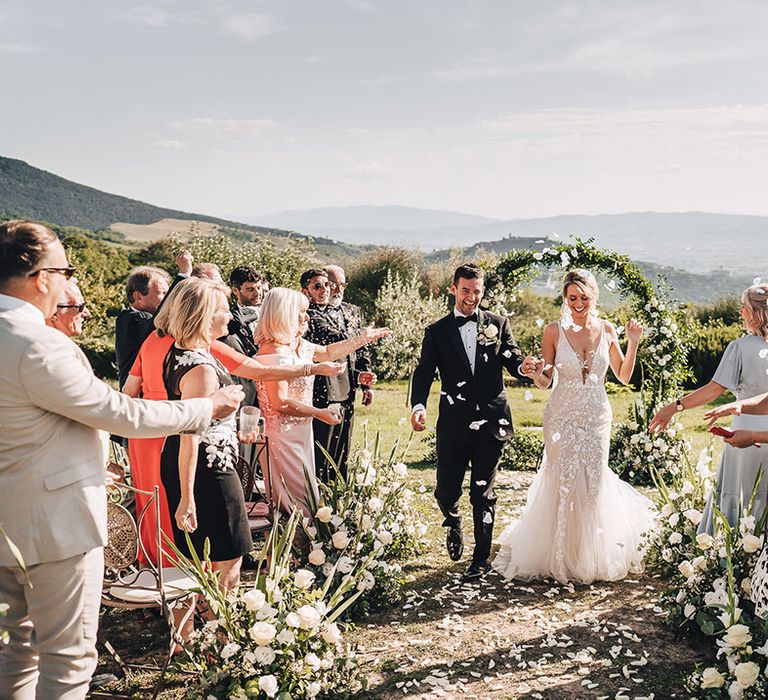 Bride in plunge lace wedding dress and trumpet styled skirt walks alongside her groom in black-tie through white petal confetti