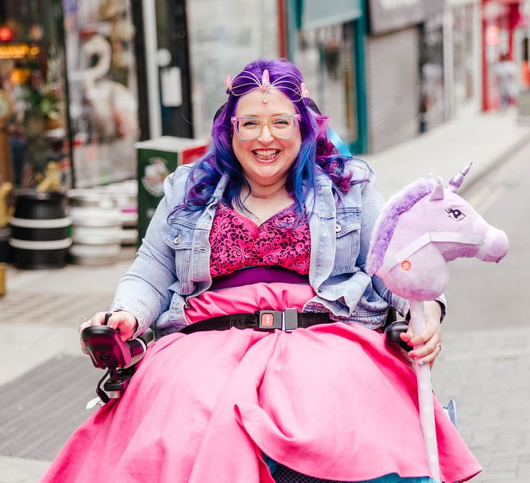 Bride in wheelchair wearing personalised denim jacket, pink wedding dress and blue fishnet tights, gemstone bridal crown, holding fluffy purple unicorn toy