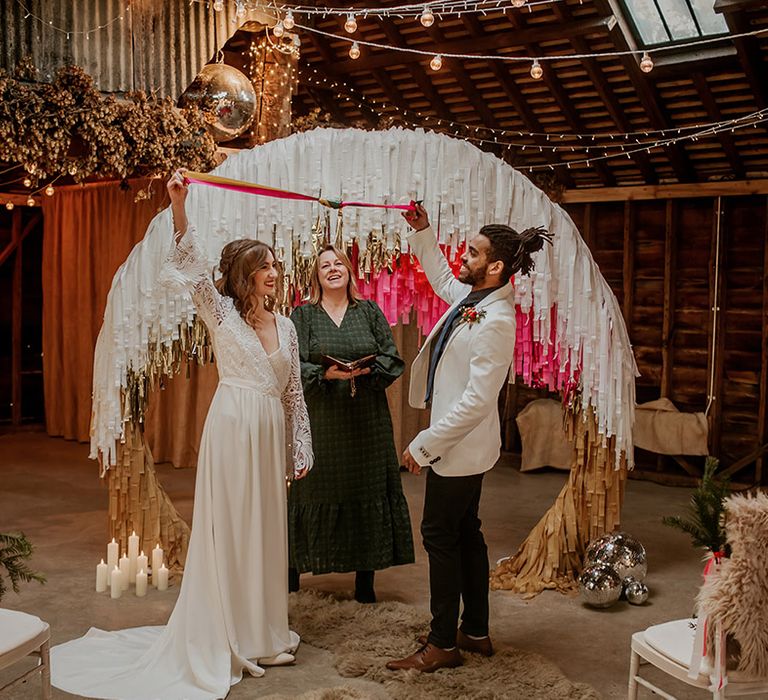 Groom in white blazer with colourful boutonniere, black shirt and black suit trousers doing handfasting ceremony with fuchsia pink ribbon and bride in long sleeve lace wedding dress with tassels standing with celebrant in dark dress and large semi-circle arch decor with white, gold and hot pink streamers 