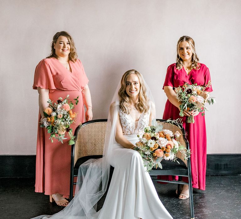 Bridesmaids wear peach and pink Rewritten Bridesmaid dresses and stand beside the bride in Mikaella Bridal wedding dress