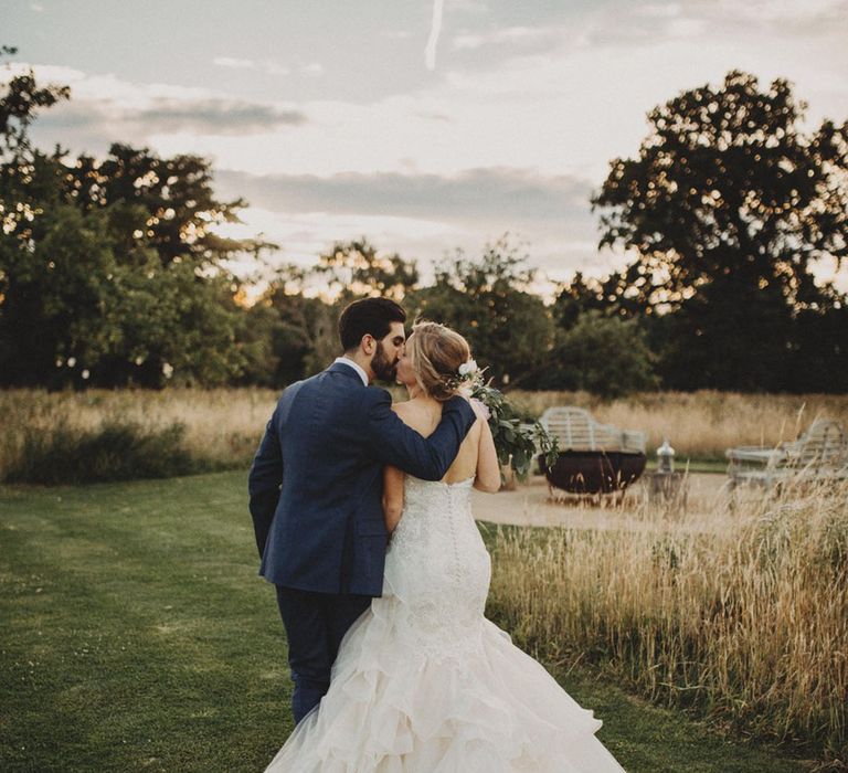 Bride in strapless button back lace mermaid wedding dress kissing the groom in a blue suit as they walk together 