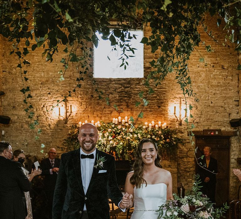 The groom in a traditional black tuxedo walks back down the aisle with the bride in a strapless mermaid wedding dress with diamanté belt accessory