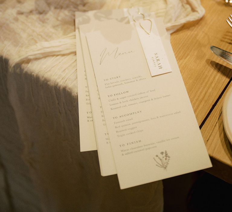 White wedding menu with gold paperclip and white place name on white table runner with small glass vase with white flower arrangements 