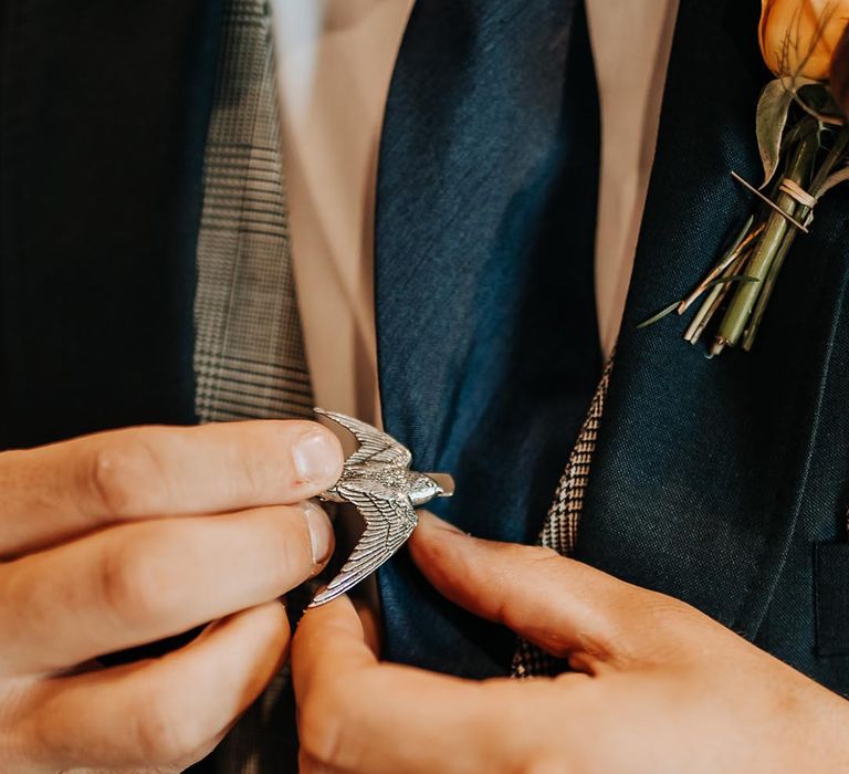 Groom attaching on a silver bird tie clip to navy tie clip 