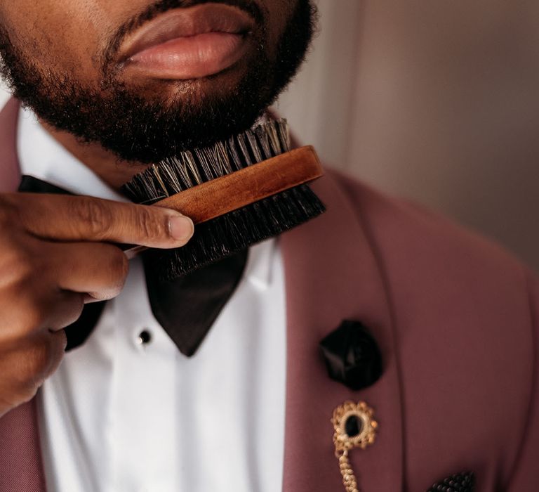 Black groom in dusky pink suit jacket with white shirt and black bow tie and gold chain accessory with bristle brush brushing his beard 