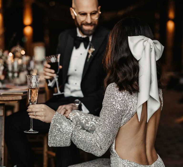 Bride in open back detail long sleeve silver sparkly wedding dress and large white satin bow bridal hair accessory talking to groom in classic sleek tuxedo with black bowtie and boutonniere 