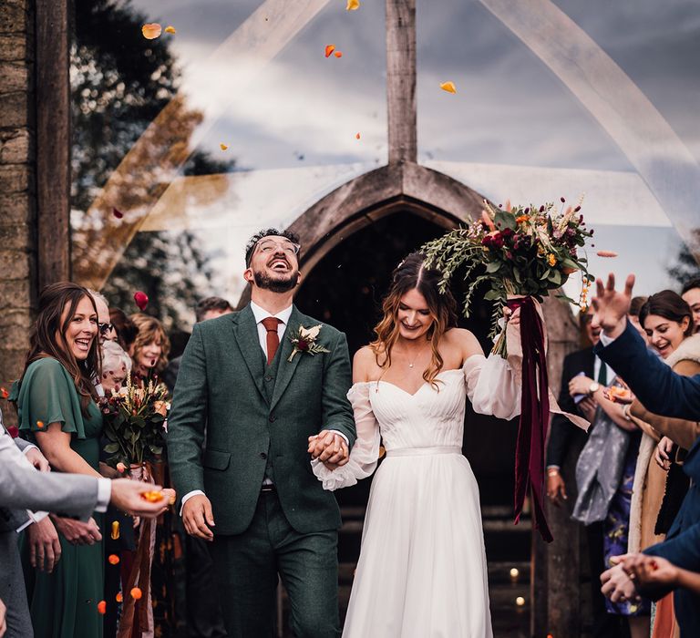 Groom in three piece dark coloured suit with dark orange tie and dried flower buttonhole walking with the bride in an off the shoulder balloon sleeve wedding dress