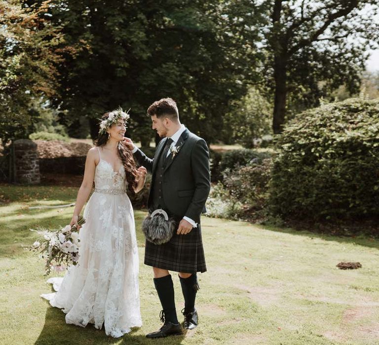 Bride wearing ethereal, celestial 3d appliqué sheer overlay wedding dress and bridal flower crown holding large white rose, peony, dried flower and foliage bridal bouquet walking with groom in dark grey blazer and waistcoat, dark tartan tie with white flower boutonniere, dark grey and blue tartan kilt and wedding sporran 