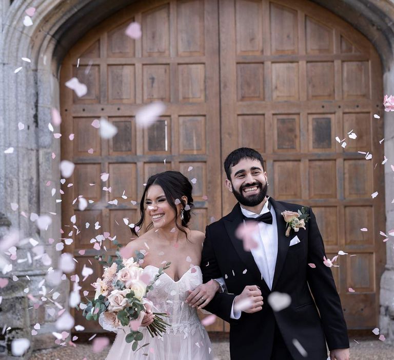 Groom in classic black tuxedo with bowtie and garden rose and eucalyptus boutonniere walking with bride in ethereal off the shoulder wedding dress with detachable puff tulle sleeves and embroidered rose 3d appliqué details doing wedding confetti exit at Pentney Abbey wedding