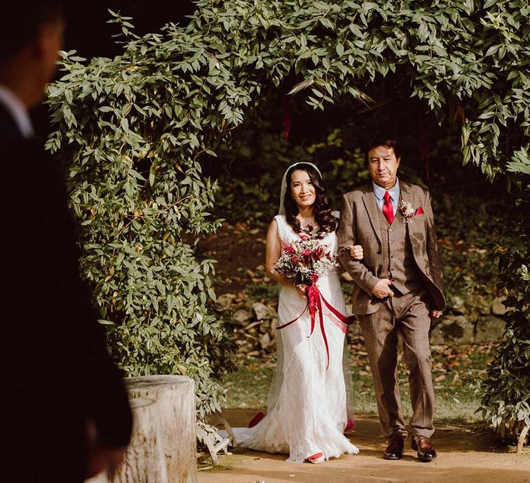 Bride in sleeveless lace wedding dress holding large red rose and dried flower bridal bouquet walking down the aisle at The Orangery Maidstone wedding venue 