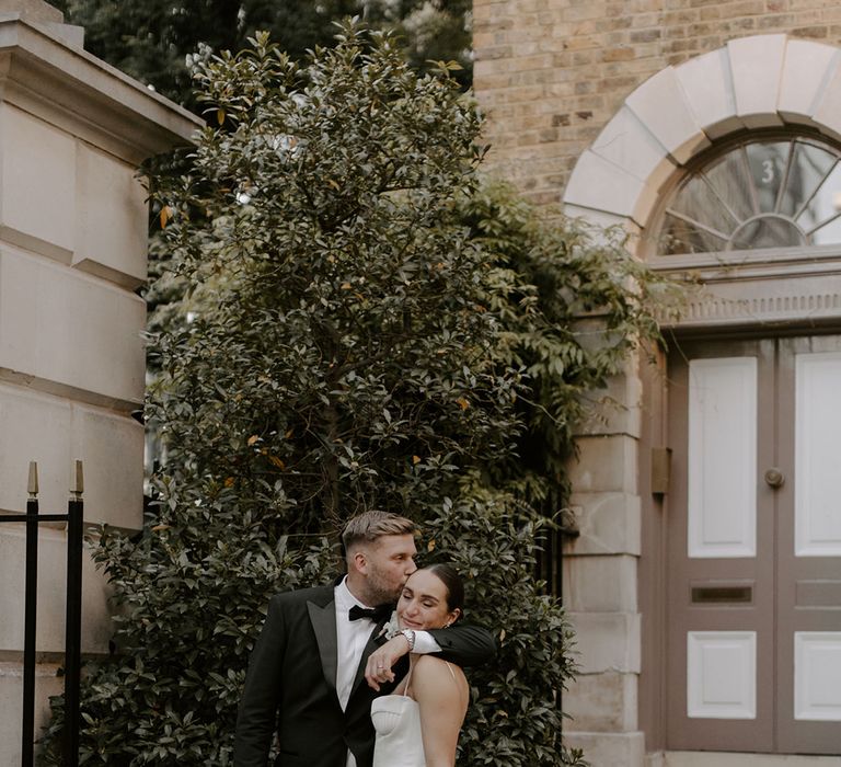 The groom affectionately kisses the bride on the forehead as they pose for their wedding photos together 