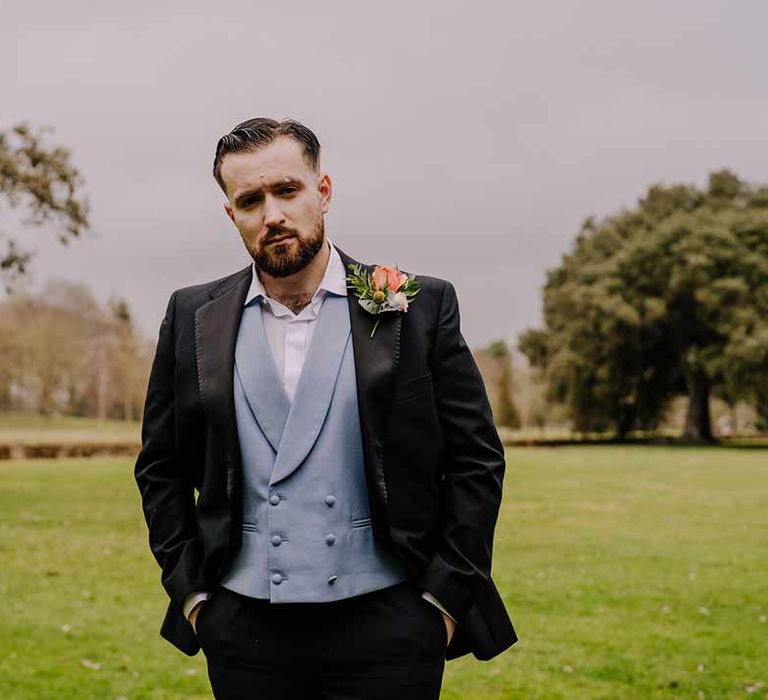 Groom wearing black blazer with pink garden rose and foliage boutonniere, cornflower blue waistcoat and black suit trousers at Braxted Park wedding venue 