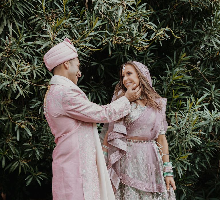 Bride in mint green and dusky pink ombre embellished lehenga posing with groom at destination wedding wearing light pink sherwani, light pink turban and cream trousers  