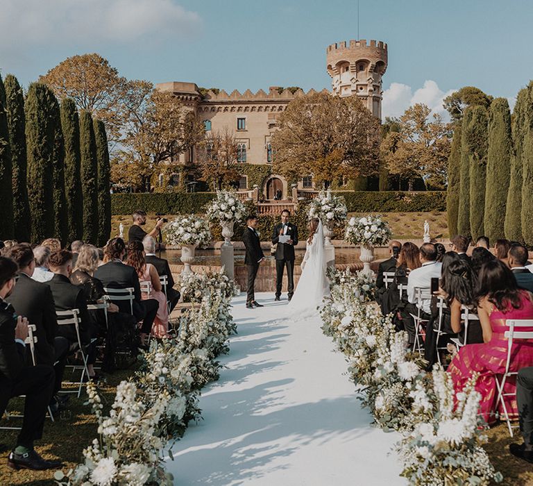 Bride and groom at the end of the aisle with carnation, eucalyptus and dried flower aisle flower decorations with 13th-century castle wedding venue in Barcelona behind them 