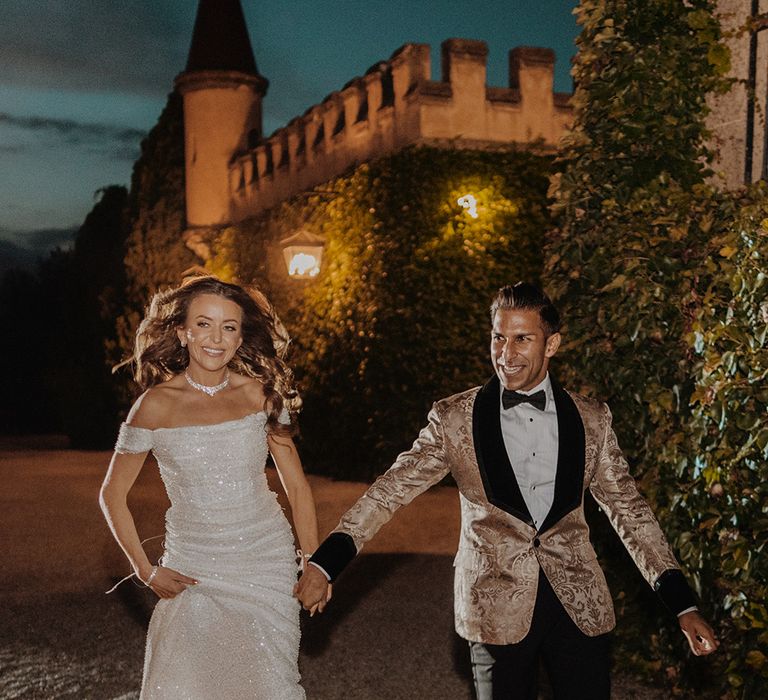 Bride in off the shoulder reception dress running with groom in gold blazer with black lining, black bowtie and black suit trousers 