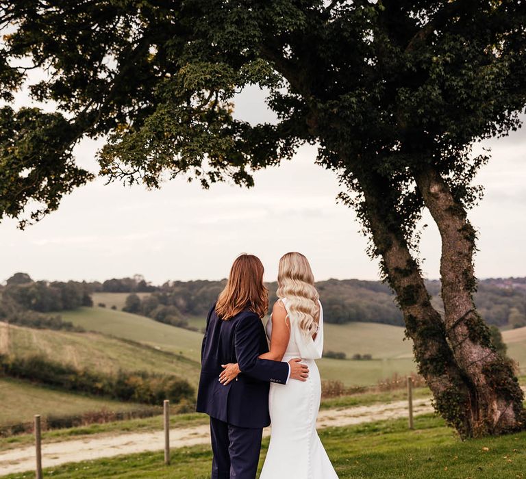 Groom in blue suit looking out at the view of the Surrey Hills with the bride in a draped back Savin London wedding dress with Hollywood curled hair 