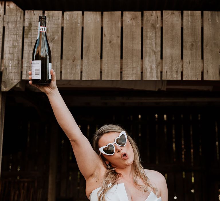 Bride in strapless Suzanne Neville wedding dress wearing white retro heart sunglasses raising a bottle of champagne in the air 