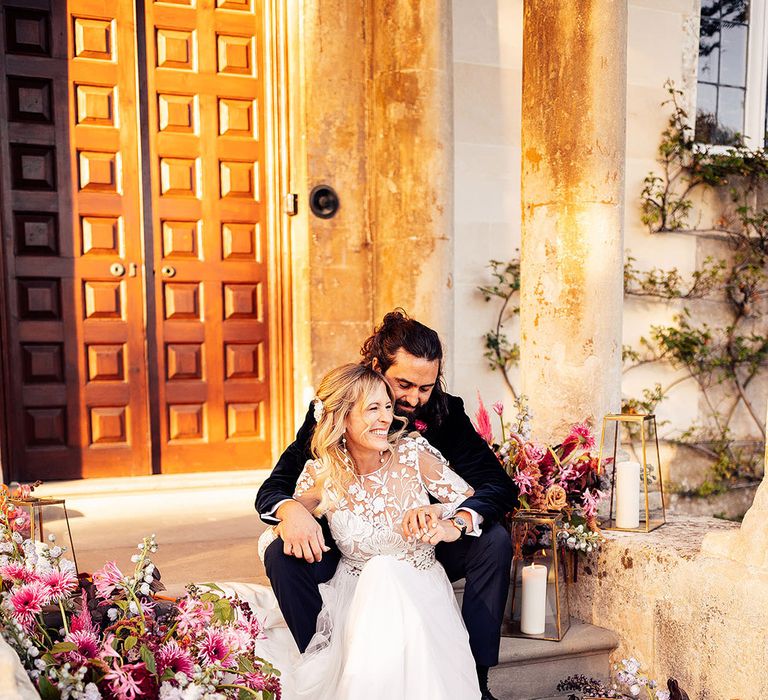 Laughing bride in luxe Margaux Tardits wedding dress sitting with groom on the steps to Elmore Court 